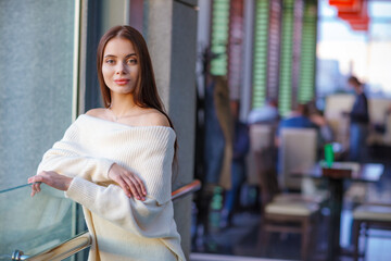 Beautiful model girl with dark long straight hair holds smiles, looking at the camera, white clothes, folded arms. Near a gray wall in an open city. Blurred details and background