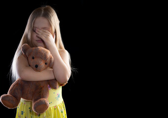 Little girl hugs a toy bear on a dark background, cries and covers her face with her hands