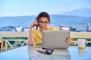 Middle aged woman working at home on a terrace with laptop and smartphone