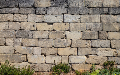 Texture, background, pattern. Ancient dry built stone brick wall. Irregular stones broken and ruined by time. Weeds grow low. Rough and dirty surface.