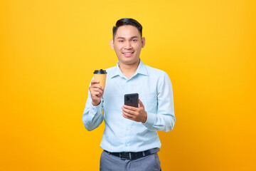 Smiling young handsome businessman holding smartphone and cup of coffee on yellow background