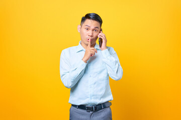 Young handsome businessman talking on smartphone and make silence gesture on yellow background