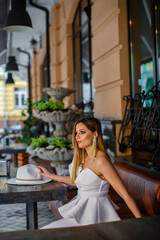 Naklejka premium Close up fashionable woman portrait of young pretty fashionable girl posing in white dress and retro hat while sitting in cafe. Fashion chic