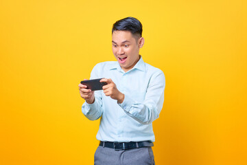 Excited young handsome businessman playing a game on mobile phone on yellow background