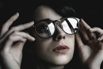Portrait of a girl with glasses. Close-up of a face looking at the camera. The girl straightens her glasses with her hands. Black background. In the lenses of glasses reflections