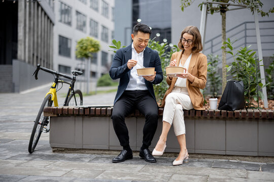 Asian Businessman And Caucasian Businesswoman Eating Food And Talking While Having Lunch At Work. Concept Of Rest And Break On Job. Smiling Business People Sitting On Bench In City