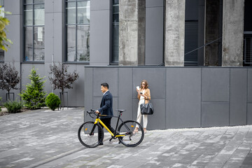 Asian businessman and caucasian businesswoman on city street. Asian guy passing by with a bicycle, woman using phone. Business people during a break outdoors. Wide view on the office building