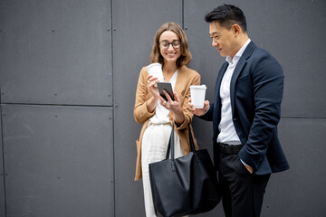 Asian businessman and caucasian businesswoman drinking coffee and talking in city. Concept of business cooperation. Idea of freelance and remote work. Woman with smartphone. Modern successful people
