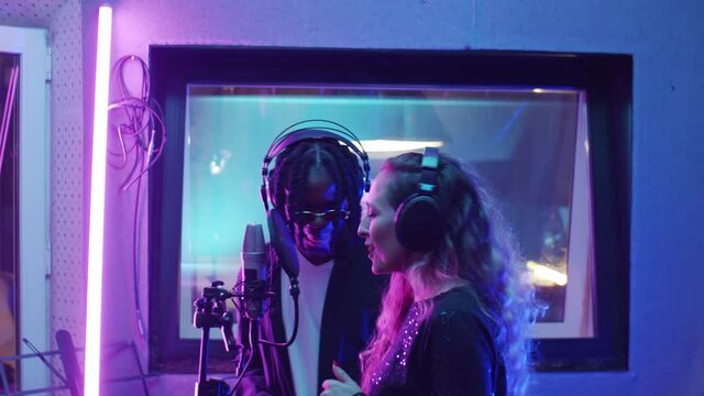 Caucasian woman and African American man in headphones standing in recording studio with neon light and singing together in microphone