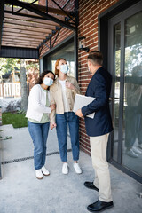 realtor holding folder near lesbian multiethnic couple in medical masks while showing new house