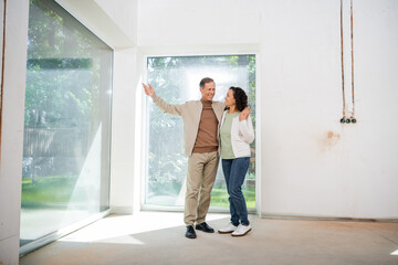 Fototapeta na wymiar happy man pointing with hand at panoramic windows while hugging wife in new house