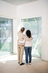 back view of husband pointing with finger at panoramic windows while hugging wife in new house