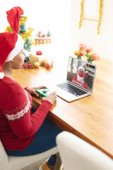African american woman in santa hat making laptop christmas video call with santa claus in face mask