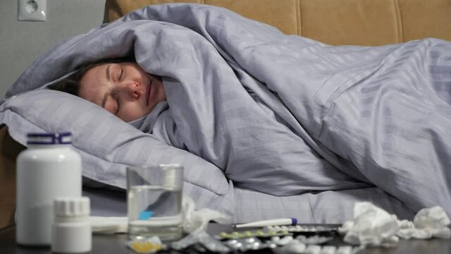 Young woman with runny nose coughs heavily lying on sofa under comfortable blanket near table with prescribed medications in living room.