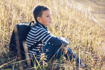 Autumn portrait of teenager boy sitting on grass. Young man with backpack resting relaxing,...