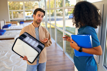An African American female salesperson consultant advises a man who is holding a mattress sample in...