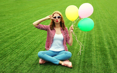 Happy young woman with bunch of balloons sitting on the grass in a summer park