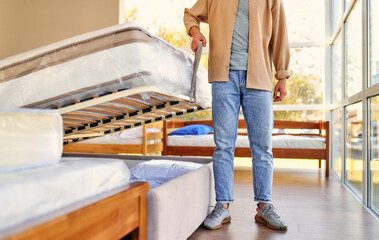 A man chooses a bed in a store, examines a frame with slats. Purchase of a mattress, bed and other sleeping accessories.
