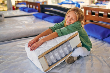 Beautiful child cute girl sitting on the bed and holding a cutaway mattress sample in the store....