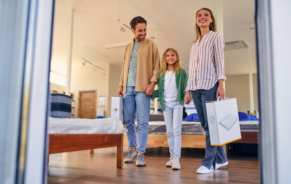 Young Happy Family With Cute Daughter Leaving Joyful Out Of The Store With Purchases Of New Pillows. Shopping For Mattresses, Beds And Other Bedding.