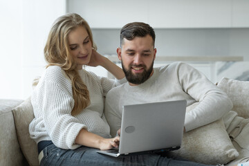 Happy young couple is using a laptop together, sitting at home on a cozy sofa, a man and a woman are looking at a computer screen