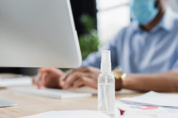 Hand sanitizer near computer and blurred african american businessman in medical mask