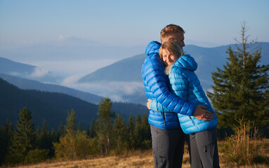 Warm embrace of husband and smiling wife against the backdrop of mountain hills covered with white fog. Tourist walking in the mountains in the morning. Concept of hiking, travelling, relationships.