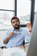 Fototapeta premium African american businessman holding eyeglasses and gesturing near blurred computer in office