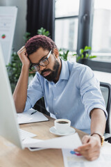 Tired african american businessman in eyeglasses holding documents near coffee and computer in office