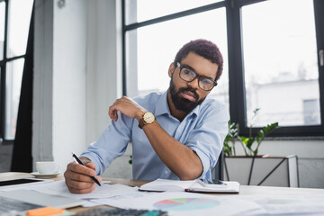 African american manager holding pen near papers and gadgets in office