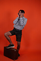 A charismatic handsome guy in a shirt and shorts stands with a suitcase on an orange background.