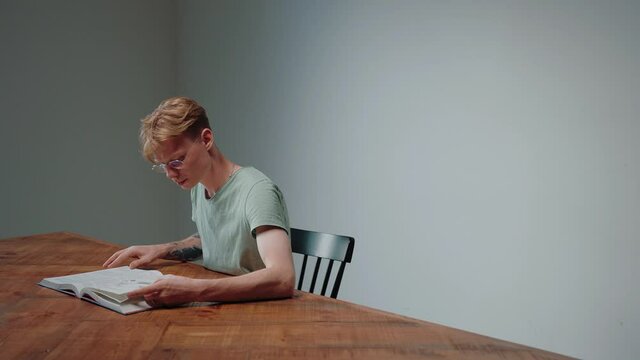 Guy in glasses studying by reading a textbook, sitting in empty grey room. Young man studying in plain grey room. Doing homework in the evening. High quality 4k footage