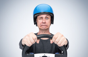 Emotionally scared man in blue motorcycle helmet looks at the road with one eye, gripping the steering wheel in his hands, on light background.