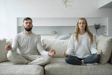 Fototapeta na wymiar Calm young family is doing yoga together, sitting in the lotus position on the sofa. Parents relax on the couch, meditating