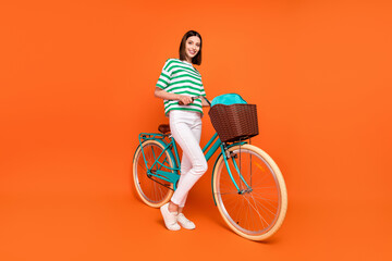 Full size photo of cute brunette young lady travel on bicycle wear t-shirt trousers sneakers isolated on orange color background