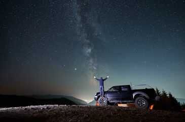 Back view of man with outstretched arms standing on rubber front wheel of black jeep in the middle of rocky mountain road and watching on night fairytale starry sky. The Milky Way over mountains.