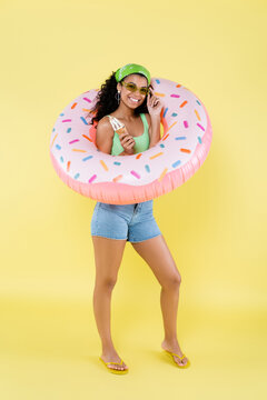 full length of joyful african american young woman standing with inflatable ring and ice cream cone on yellow