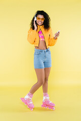 full length of african american young woman in pink roller skates and headphones using smartphone on yellow
