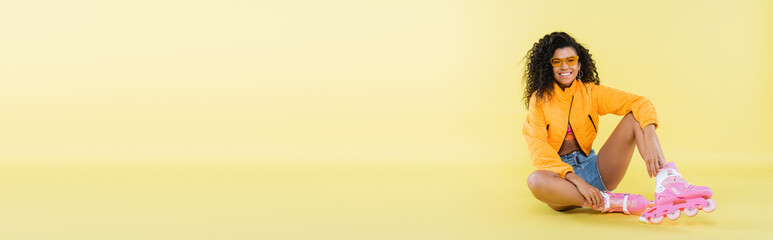 full length of smiling african american young woman in pink roller skates sitting on yellow, banner