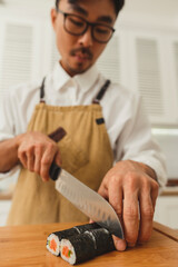 Sushi master makes sushi rolls. Portrait of asian man chef in uniform slicing finished rolls into pieces on wooden plate