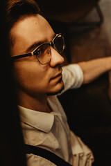 A young man sits in a leather armchair and ponders important matters. He is wearing a white shirt, black trousers with suspenders and glasses. Stylish hairstyle on the head