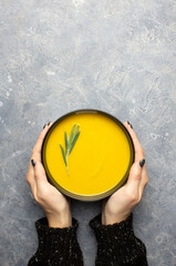 Pumpkin soup in female hands on a gray concrete background with copy space. Hands of a girl in a black sweater, a sprig of rosemary in the soup