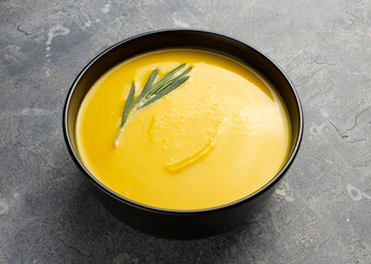 Pumpkin soup in black bowl with rosemary sprig on gray concrete surface