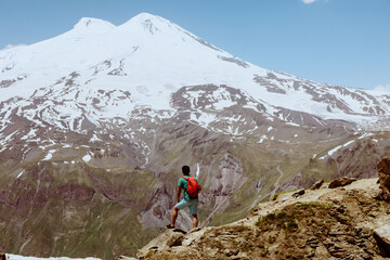 Fototapeta premium Man with red backpack stand on rocky mountain and look at snowy peak of Elbrus.
