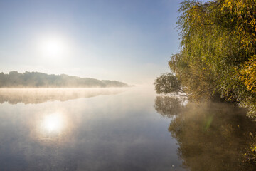 Autumn landscape in the early morning on the river. Misty water surface. Yellow leaves on trees and bushes are illuminated by the rays of the rising sun. Dawn on a cold autumn morning.