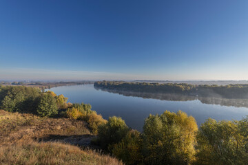 Autumn landscape in the early morning overlooking the river. A wide river and endless expanses of fields. Yellow leaves on trees and bushes are illuminated by the rays of the rising sun.