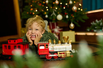 Happy curly-haired boy is playing with a magic toy train at the Christmas tree. The concept of...