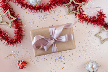Birthday or New Year's eve present box in kraft paper with pink ribbon on beige background. Xmas composition of gold stars, silver balls, glitter confetti and red tinsel. Flat lay. Giving love concept