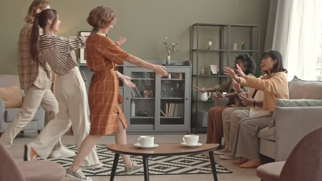 Full shot of three multiethnic young women sitting on couch in living room in apartment, Caucasian female and male friends entering and hugging each other