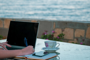 Woman remotely working from laptop and writing notes seaside on holiday. 